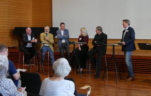 Quelle: Katholische Akademie der Erzdiözese Freiburg Vier Männer und eine Frau stehen an Stehtischen in der Aula und diskutieren.